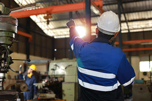 Rear view technician engineer in protective suit standing and using walkie talkie radio while controlling or maintenance operation work lathe metal machine at heavy industry manufacturing factory