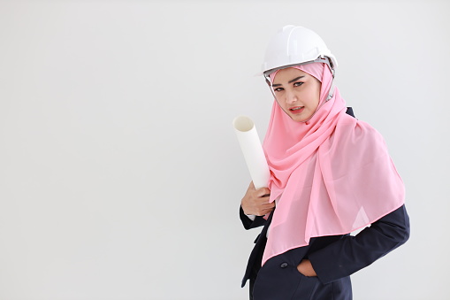 Smart muslim young asian woman wearing blue suit smiling confident holding blueprint in studio. Isolated white background portrait with beautiful face girl with pink hijab. Advertisement concept.