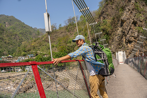 Young male Indian hiker with backpack looking at river while standing on footbridge during summer vacation