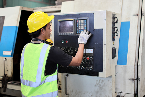 Technician engineer man in protective uniform standing and using computer  monitor while controlling operation or checking industry machine process with hardhat at heavy industry manufacturing factory