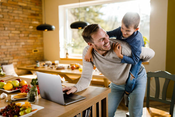 istock Happy single father using laptop and piggybacking his son in the kitchen. 1495019047