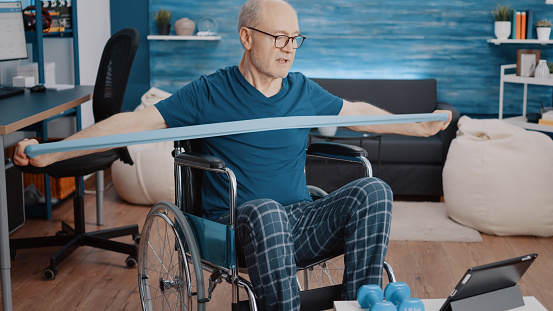 Man sitting in wheelchair and doing exercise with resistance band, looking at video of workout lesson on tablet. Aged person with disability using elastic belt, watching training program