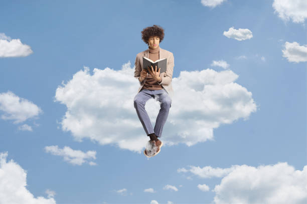 Elegant young man with curly hair sitting on a cloud and reading a book stock photo