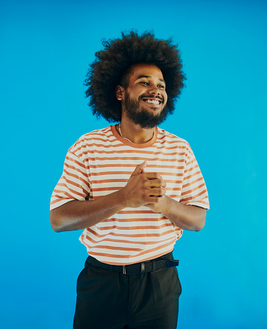Young black man with a afro hairstyle, beard and mustache holding his hands together whilst looking away from the camera smiling with dental braces and wearing a striped t-shirt, black pants and a belt. Stock photo