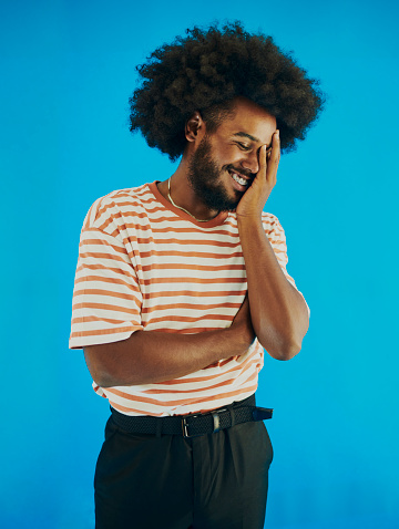 Young black man with a afro hairstyle, beard and a mustache holding his face in his hand smiling with his eyes closed wearing a striped t-shirt, black pants and belt, stock photo