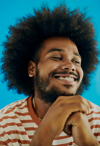 Headshot of a young black man with a afro hairstyle, beard and a mustache looking to the side whilst his hand is under his chin whilst smiling with dental braces wearing a striped t-shirt, stock photo