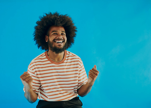 Happy young black man wearing a striped t-shirt with an afro hairstyle, punching the air looking up, shot against a blue background. Copy space . Stock photo