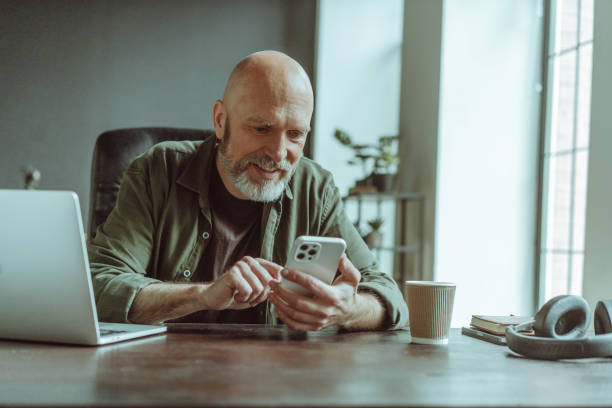 Mature old man comfortably seated home, actively engaged in texting message on phone. Desk adorned with laptop, headphones, and cup of coffee, indicating setup conducive to work and relaxation. . High quality photo anciano maduro cómodamente sentado en casa, participando activamente en mensajes de texto por teléfono. escritorio adornado con computadora portátil, auriculares y taza de café, lo que indica una configuración propicia para el trabajo y la relajación