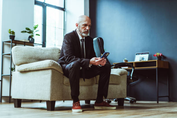 istock Old businessman dressed in black suit can be seen either texting on phone or browsing internet. Mature man, with distinguished silver beard, sits comfortably in chair within stylish loft office. 1495032153