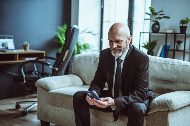 istock Senior businessman dressed in black suit, sporting warm smile, while browsing internet on phone. Mature man, with distinguished silver beard, seen resting on sofa in loft office. 1495032217