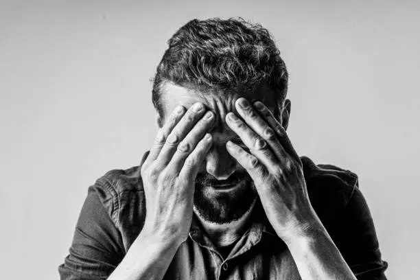 Photo of Black and white photo of a man looking extremely sad and depressed. He sitting on white background and has face covered with both hands, showing the despair and hopelessness he feels.