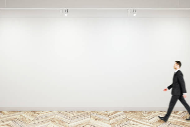 istock Man walking past blank white wall in modern abstract gallery interior, with empty space for poster or banner, mockup 1495036413