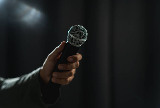 istock Confident successful speaker man talking on stage with spotlight strike through the darkness at corporate business event. Public speaker giving talk at conference hall. Stand up comedian. 1495043887