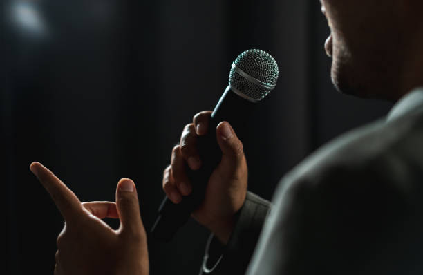 istock Confident successful speaker man talking on stage with spotlight strike through the darkness at corporate business event. Public speaker giving talk at conference hall. Stand up comedian. 1495043888
