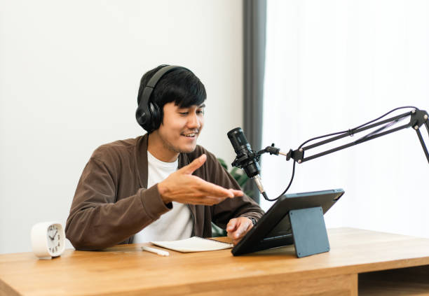 istock Young asian man host streaming podcast with condenser microphone work on tablet at small broadcast home studio. Content creator blogger recording voice over radio interview guest conversation 1495046893