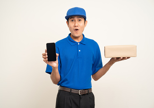 Asian man in blue uniform standing holding box parcel cardboard and smartphone on isolated white background. Male service worker with cell phone. Delivery courier shipping service