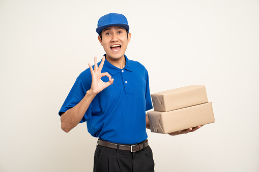 Asian man in blue uniform standing holding box parcel cardboard and smartphone on isolated white background. Male service worker with cell phone. Delivery courier shipping service