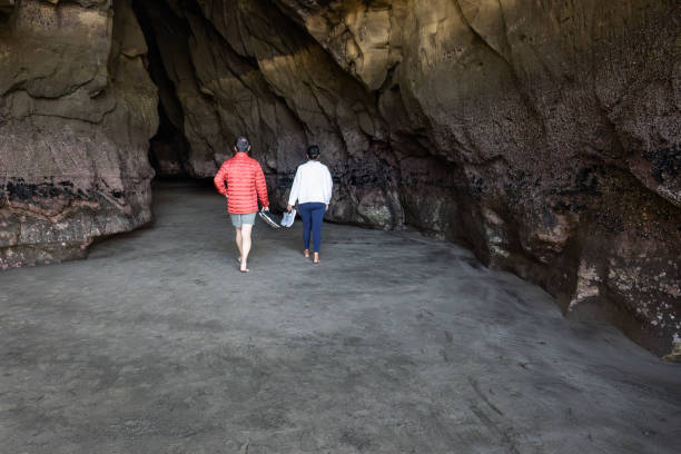 junges paar hält schuhe in den händen und geht barfuß in richtung einer höhle am strand von muriwai. auckland. - activity asia atmosphere beach stock-fotos und bilder