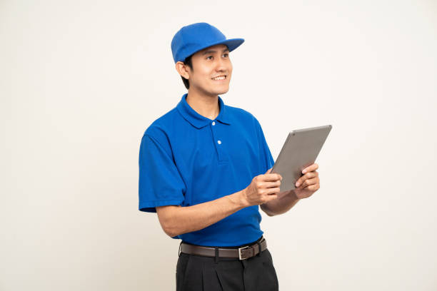 istock Asian man in blue uniform standing holding digital tablet computer on isolated white background. Male service worker with cell phone. Delivery courier shipping service 1495048061