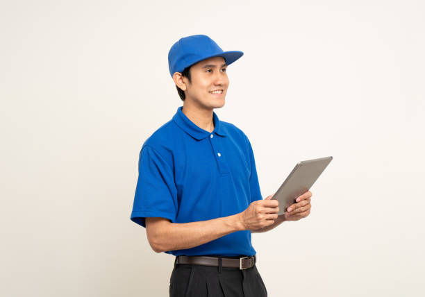 istock Asian man in blue uniform standing holding digital tablet computer on isolated white background. Male service worker with cell phone. Delivery courier shipping service 1495048065