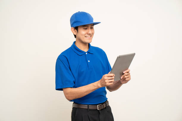 istock Asian man in blue uniform standing holding digital tablet computer on isolated white background. Male service worker with cell phone. Delivery courier shipping service 1495048979