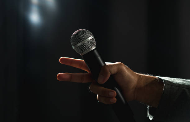istock Confident successful speaker man talking on stage with spotlight strike through the darkness at corporate business event. Public speaker giving talk at conference hall. Stand up comedian. 1495049035