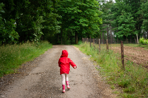 Rear view of little girl in red jacket and rubber boots running in public park during springtime