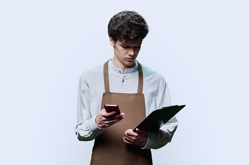 Portrait of young confident guy in apron with paper clipboard, smartphone in his hands, looking at screen of phone on white studio background. Service ordering, online order, staff occupation concept