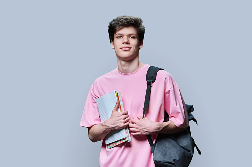 Young guy college student with backpack holding notebooks textbooks, in pink t-shirt looking at camera on grey studio background. Education, studying, learning concept