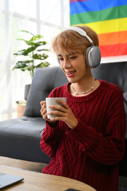 istock Pretty young gay man wearing headphone drinking coffee watching videos, browsing internet on laptop at home. 1495055568