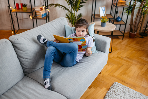 Beautiful little girl reading a book on the sofa in the living room.