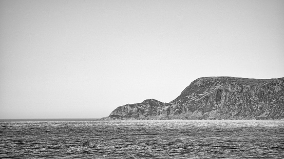 View from the sea to the West Cape in Norway in sunshine in black and white. Waves in the foreground, rocks in the background. Westernmost point of Europe mainland.