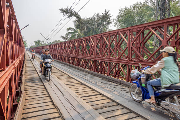 The Old French Bridge, Luang Prabang, Laos - March 18th 2023:  Motorcycles on the old wooden bridge which is from the time of the French occupation and are crossing the Nam Pak River in the former capital of Laos