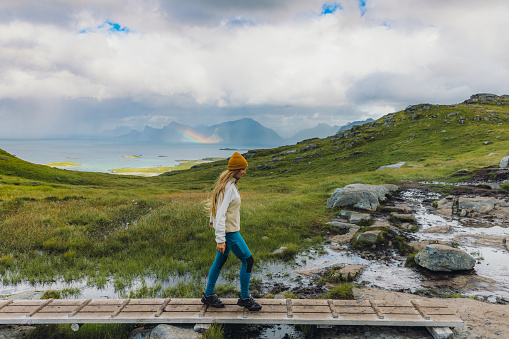 Woman traveler in yellow hat hiking in the mountains on the wooden footpath with a scenic view of the rainbow above the ocean on Lofoten, northern Norway