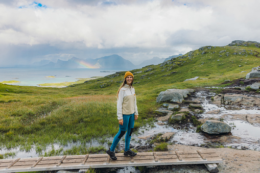 Woman traveler in yellow hat hiking in the mountains on the wooden footpath with a scenic view of the rainbow above the ocean on Lofoten, northern Norway