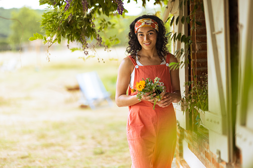 A front view of a young woman who is stood outside of a French villa with a bunch of wild flowers which she has picked. She is smiling and looking into the camera. She is wearing bright orange pyjamas.