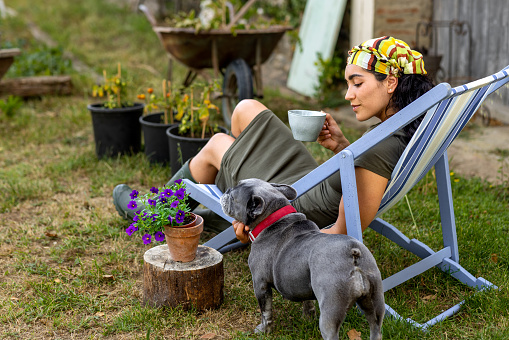 an over the shoulder view of a woman who is sitting outdoors in a garden enjoying a serene moment with a French bulldog. She is sitting in a painted deckchair in the garden of the home she is staying in Toulouse in the south of France.