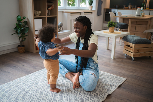 Woman Black ethnicity holding her baby boy hands, while he learning how to walk, in their home