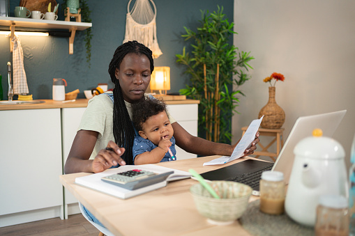 Worried single mother of Black ethnicity, analyzing and calculating her home finances, while her baby boy sitting in her lap