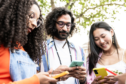 Multiethnic group of friends texting on their smartphones in the street.Three diverse young people smiling and using their phones outside.