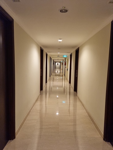 Picture of long corridor with clean marble floor, many closed doors along side, bright lighting and brown doors