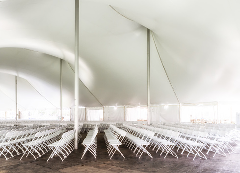 View inside a large event white tent with its billowing roof held up with poles; and rows of white folding chairs. High key lighting.