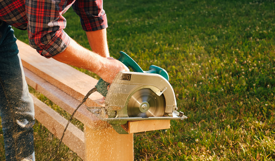 A male carpenter works with a circular saw on the lawn. Close-up view on hands of a working tool. Contrast sunlight with glare scattered in flying sawdust.