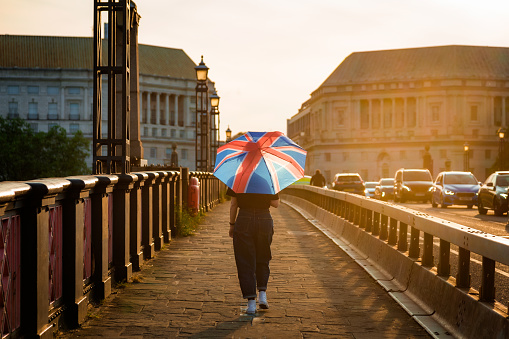 Color image depicting the rear view of an unrecognisable woman holding an umbrella with the design of the British union flag on it. It is a sunny day and the woman is using the umbrella for protection from the sun. She is walking across Lambeth bridge as the setting sun covers the scene in golden light.