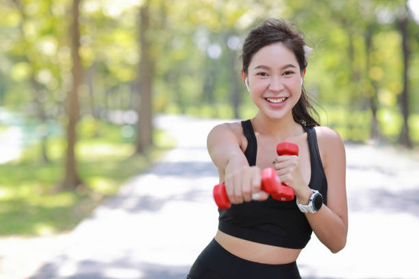 schöne asiatische fitnesssportlerin, die hantelkrafttraining nach dem morgendlichen lauf im freien hebt. glücklich lächelnde sportlerin, die unter dem baum im park trainiert, während sie in die kamera starrt - fashion model beautiful power action stock-fotos und bilder