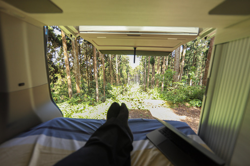 A man relaxing in a camper van, looking through the open push back door to the alpine woods, first person view, and carrying a laptop freelancer