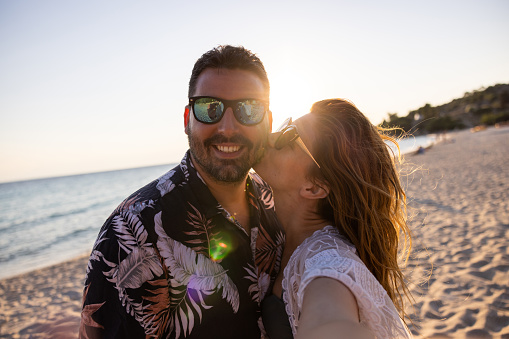 Young Caucasian woman kissing her boyfriend while taking selfie with him on the beach during sunset