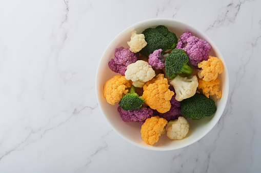 Colorfu cauliflower. Cauliflower cut into small pieces white bowl on light stone marble concrete background. Purple, yellow, white and green color cabbages. Agricultural harvest. Mock up. Top view.