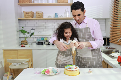 Happy lovely asian single dad and cute daughter in apron having fun with teaching and decorating homemade cake in the kitchen. Family lifestyle cooking with education concept.