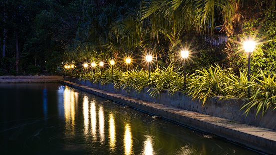 A view of the yellow light from the rows of lampposts decorated with gardens beside the pond and the light reflecting on the water in the early morning at a temple in rural Thailand.